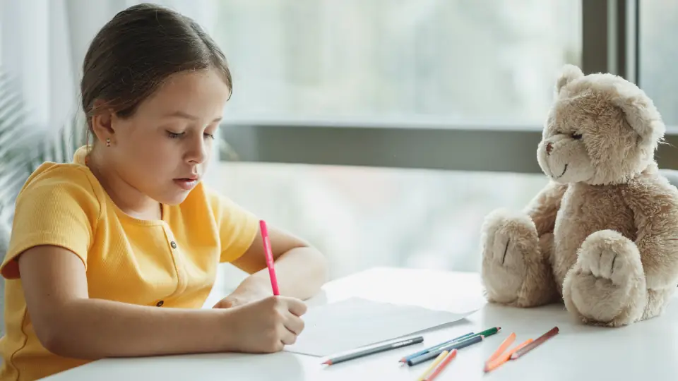 Young child drawing at a table with a teddy bear